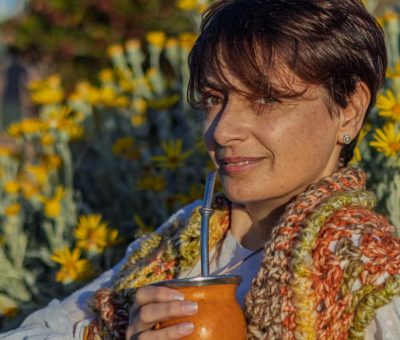 Woman with handcrafted scarf sipping yerba mate outdoors in Montevideo. Vibrant yellow flowers in full bloom.